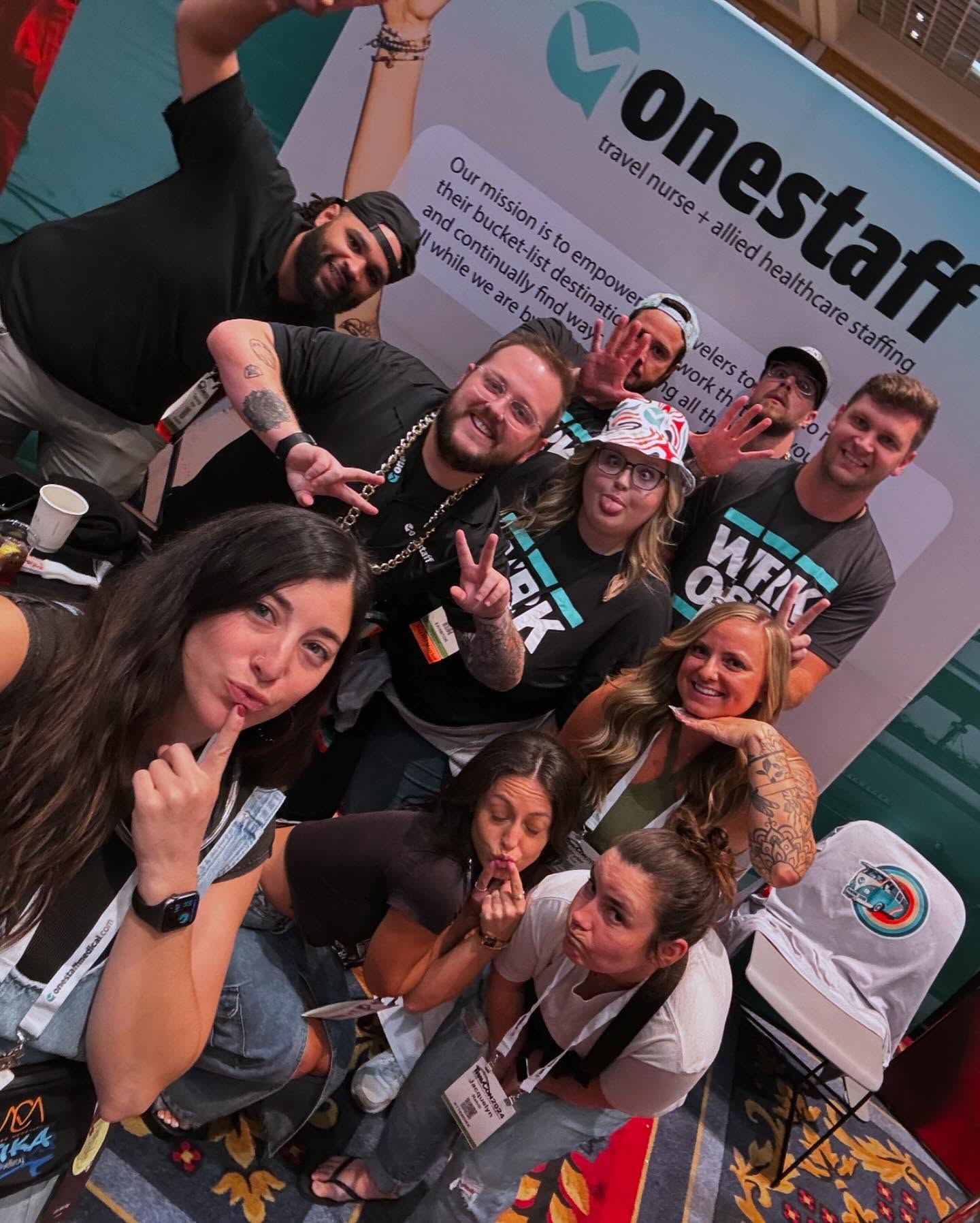 Big group selfie of the OneStaff crew and attendees throwing peace signs at the booth