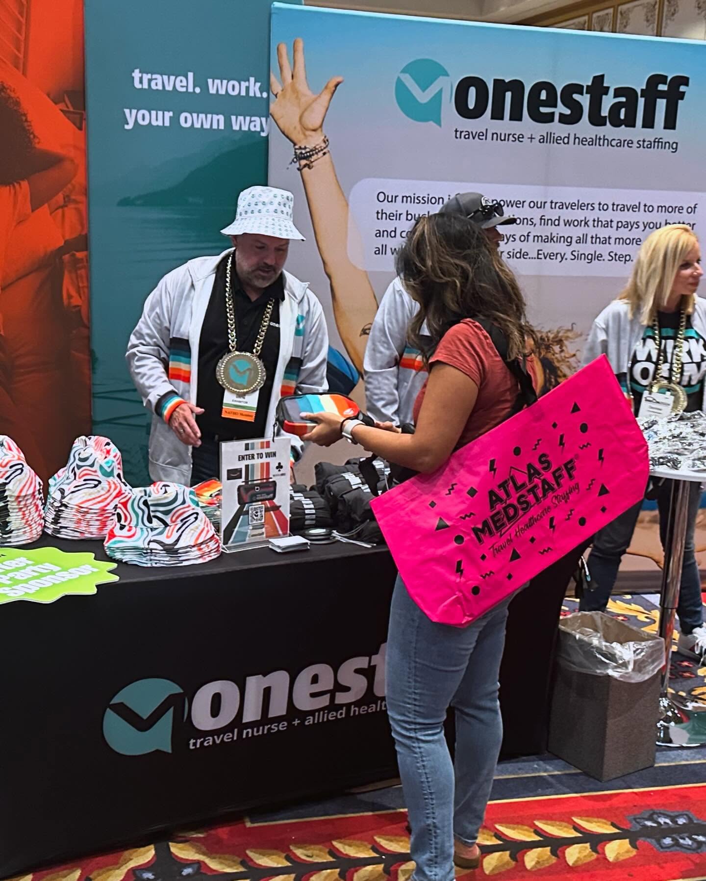 OneStaff recruiter in a champ chain and reversible bucket cap at the TravCon booth — attendee browsing the giveaway display loaded with bucket caps, fanny packs, and the enter-to-win sign