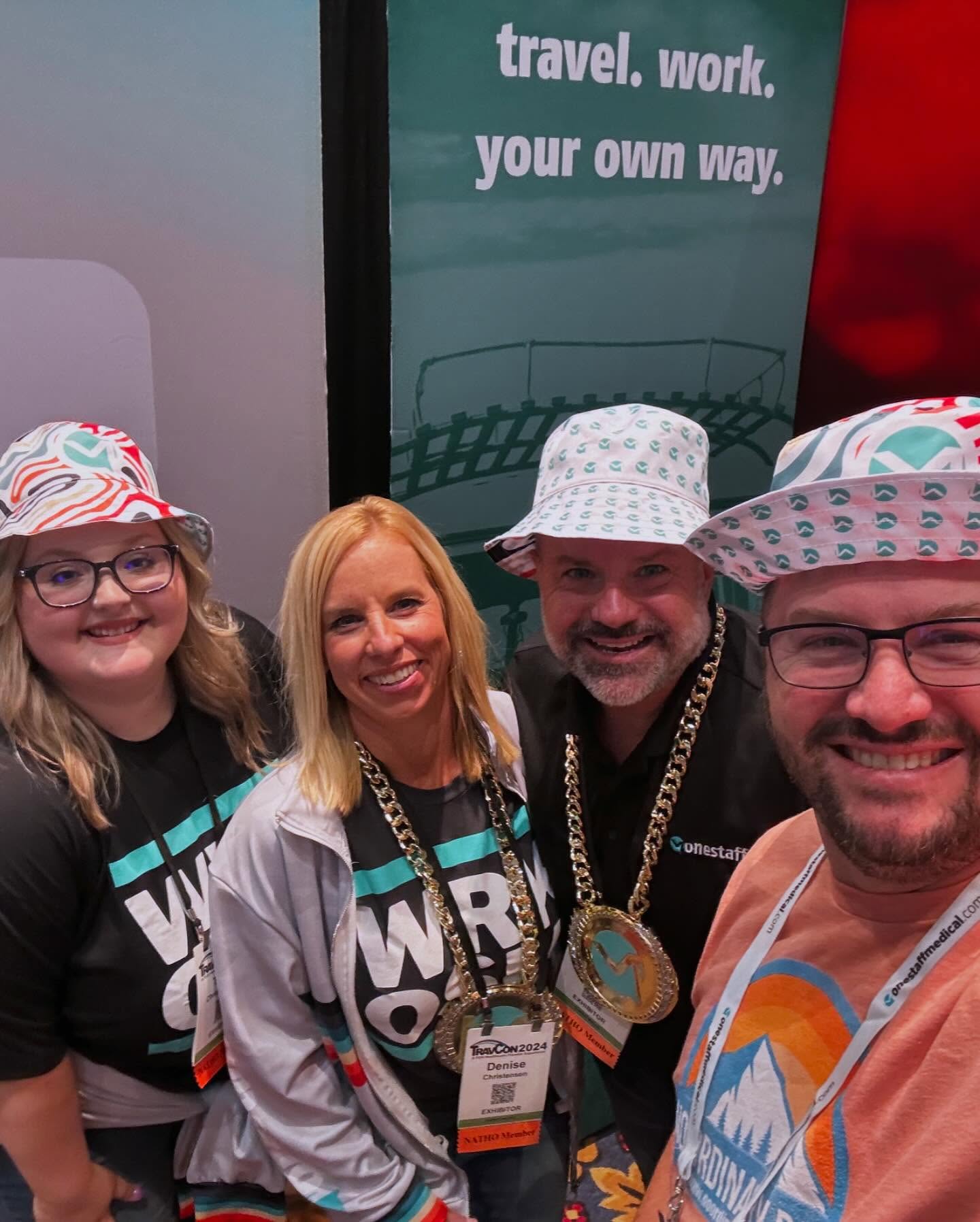 OneStaff Medical team and partners in custom bucket hats and champ chains at the TravCon booth — 'travel. work. your own way.' banner behind them
